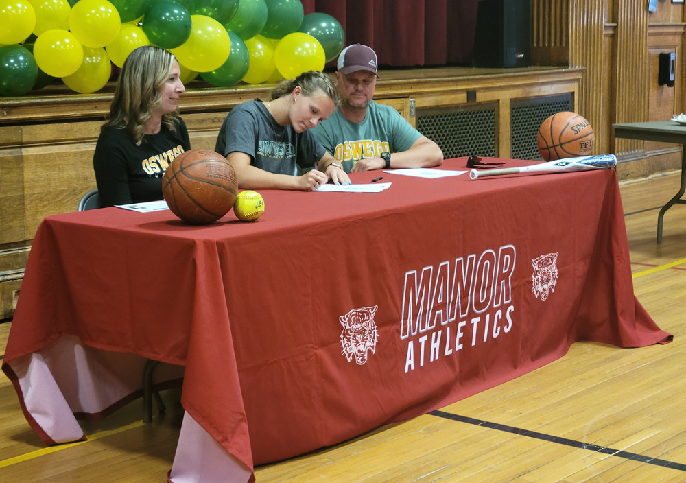 A student sitting at a table covered with a Manor athletics table cloth and green and yellow balloons behind her signs a paper as two adults sit on either side