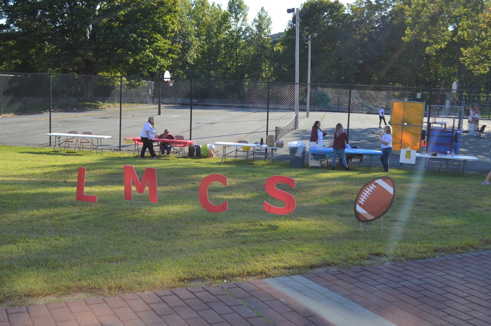 Tables are set up along the fence to the basketball court and signs with LMCS and a football are in the grass in the foreground