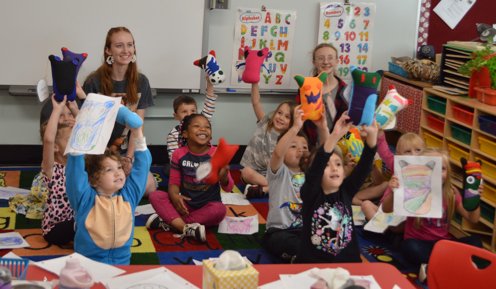 Students hold up their monster dolls and drawings in a classroom