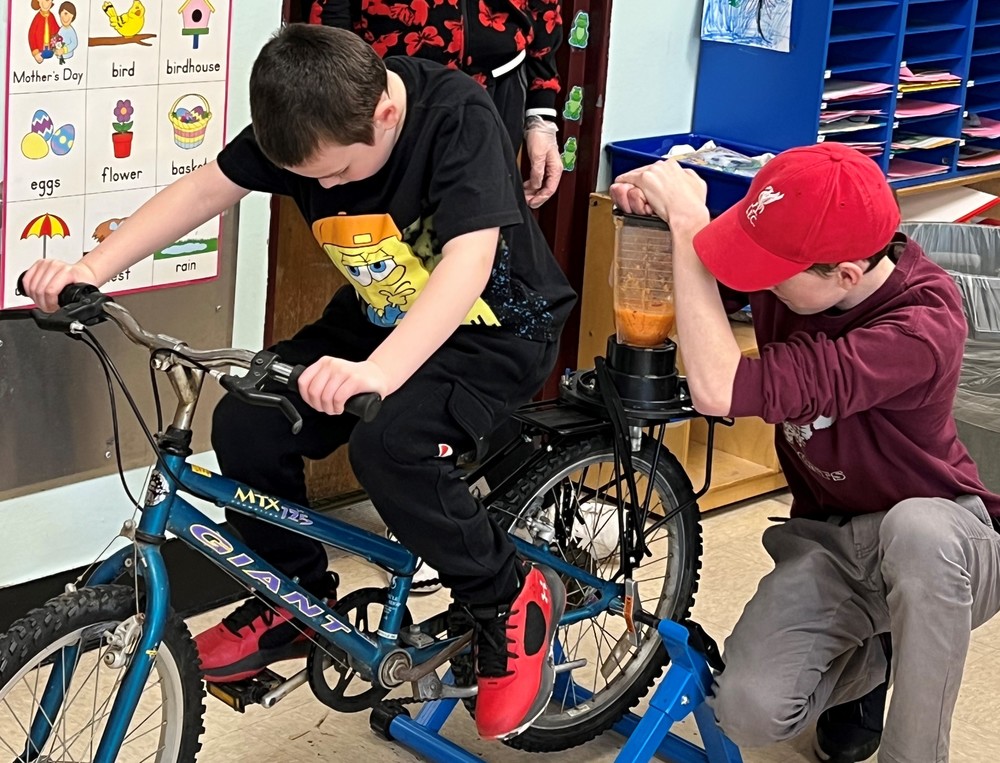 An older student holds the top of a blender as a younger student pedals the blender bike