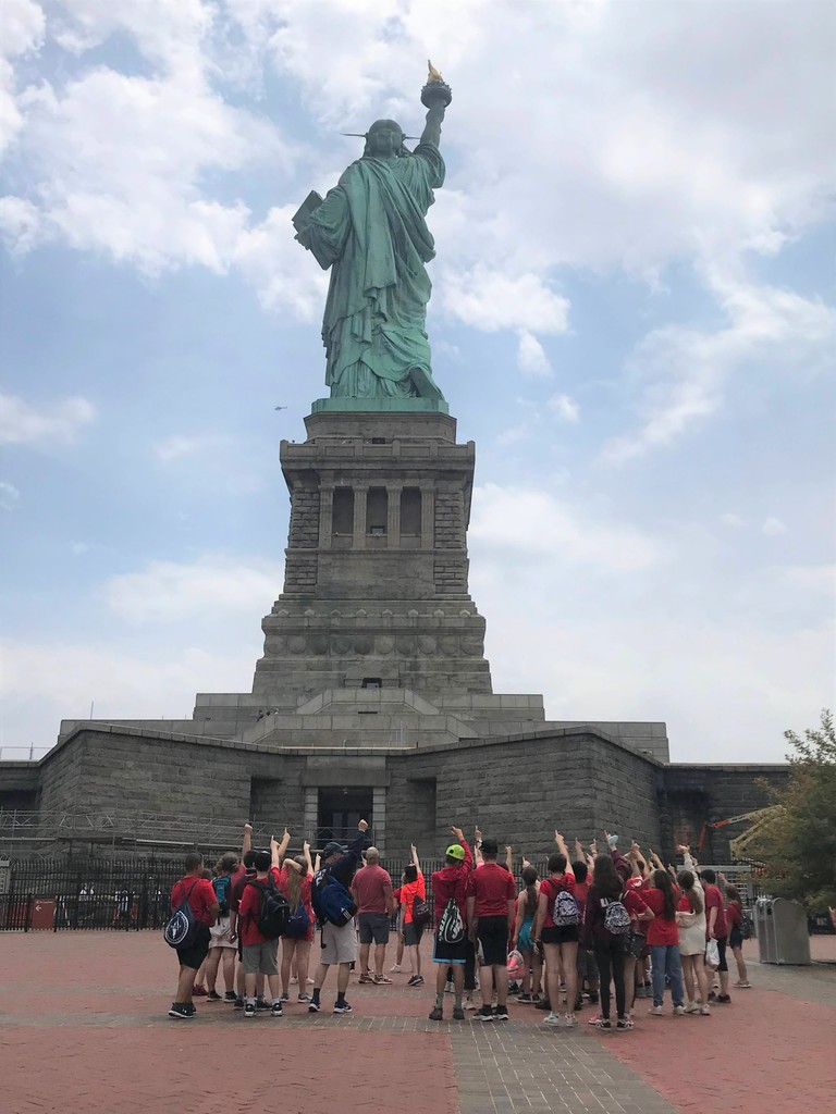 The statue of liberty is seen in the background as a group of kids hold up  their left arms  at the base.