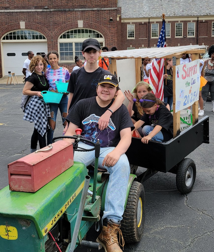 A teen sits on a garden tractor that is pulling a cart with two girls on it as other teens stand near