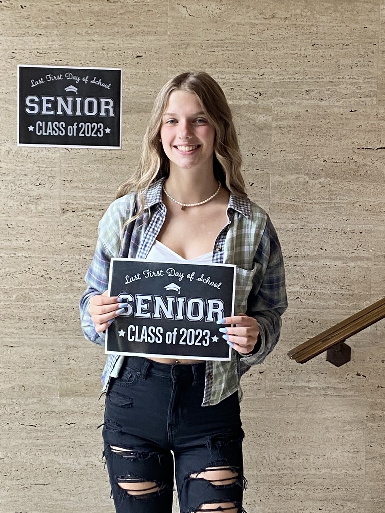 A student holds a black sign with white letters that reads Last first day of School, senior, Class of 2023 in front of tan background with the same sign  at the top left portion of the picture
