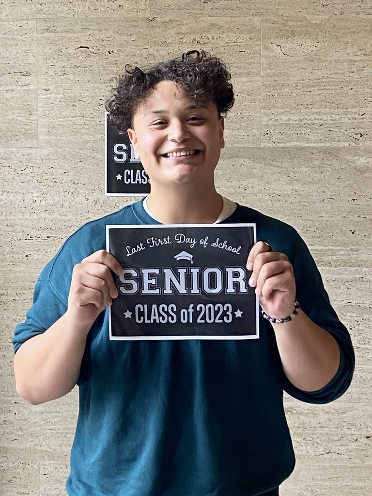 A student in a blue shirt holds a black sign with white letters that reads "Last first day of school Seniors Class of 2023"