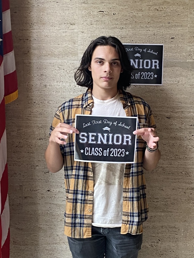 A student with a while shirt and yellow and blue plaid shirt over it and black pants holds a black sign with white letters that reads "Last first day of school Seniors Class of 2023"
