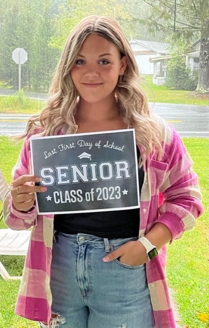 A student in a black shirt with a pink plaid shirt over it and blue jeans holds a black sign with white letters that reads "Last first day of school Seniors Class of 2023"