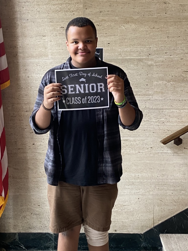 A student wearing a black shirt and a black and gray plaid shirt over it and brown shorts) holds a black sign with white letters that reads "Last first day of school Seniors Class of 2023"