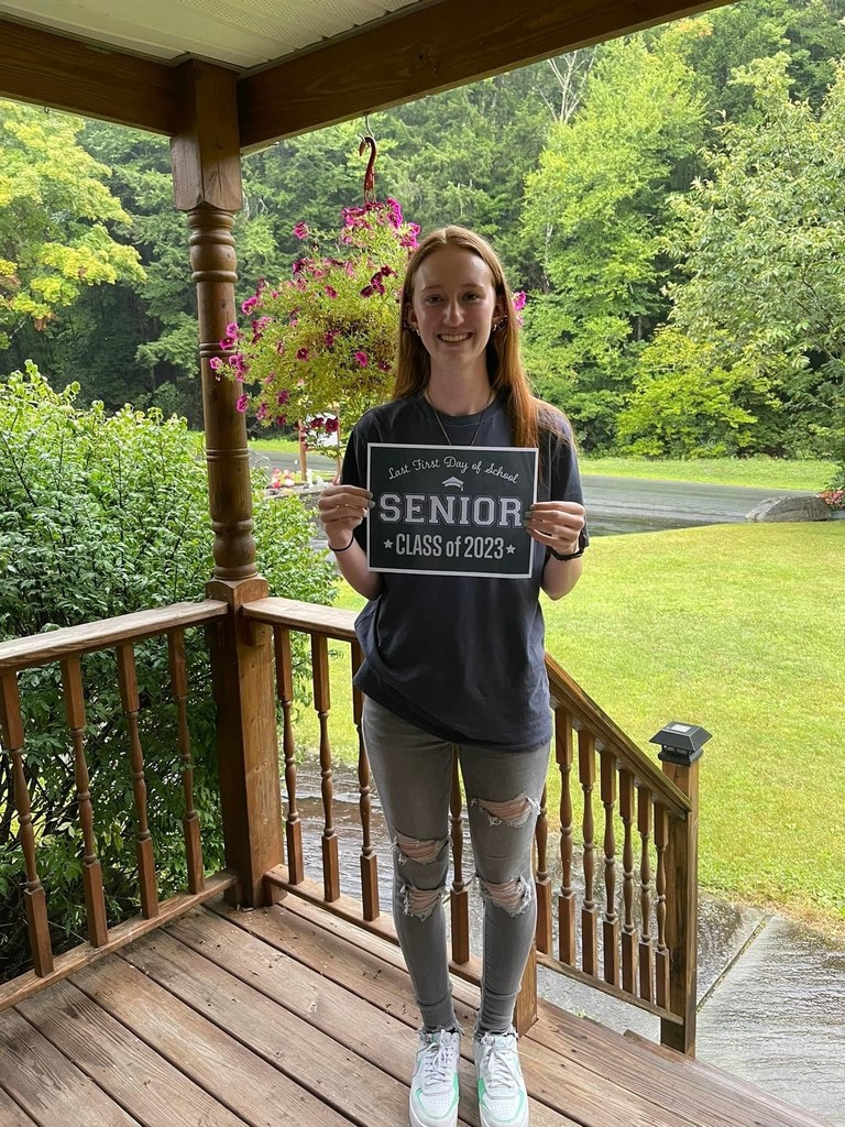 A student in a black shirt and gray ripped jeans standing on a porch with green grass and trees in the background  holds a black sign with white letters that reads "Last first day of school Seniors Class of 2023"