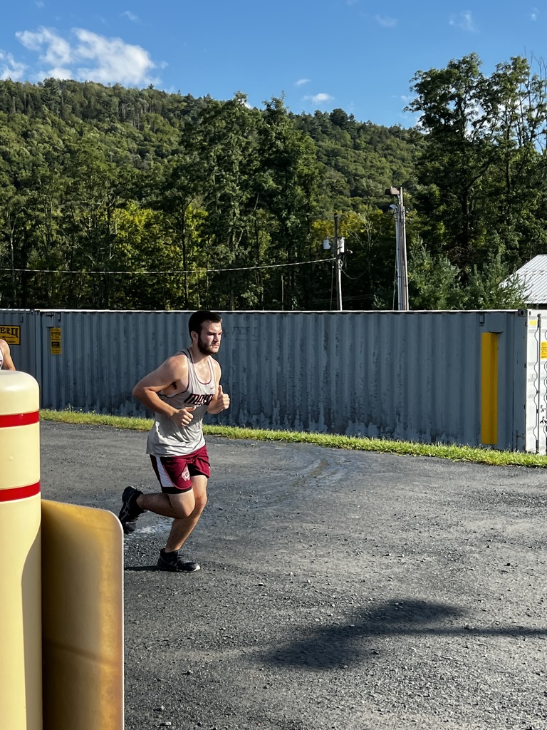 A runner in a gray shirt and maroon shorts runs on pavement with a gray shipping container behind him.