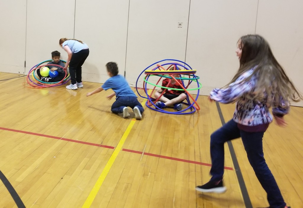 a student sits surrounded by hoops and balls as another student helps, while another student sits in a cage made of hops and students on their needs and running are nearby