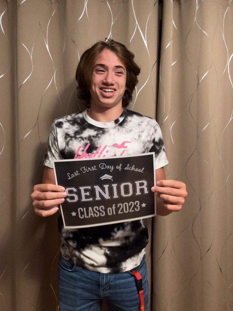 A student in a black, white and pink shirt and jeans holds a black sign with white letters that reads "Last first day of school Seniors Class of 2023"