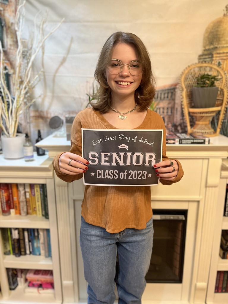 A student in a tan shirt and jeans holds a black sign with white letters that reads "Last first day of school Seniors Class of 2023"