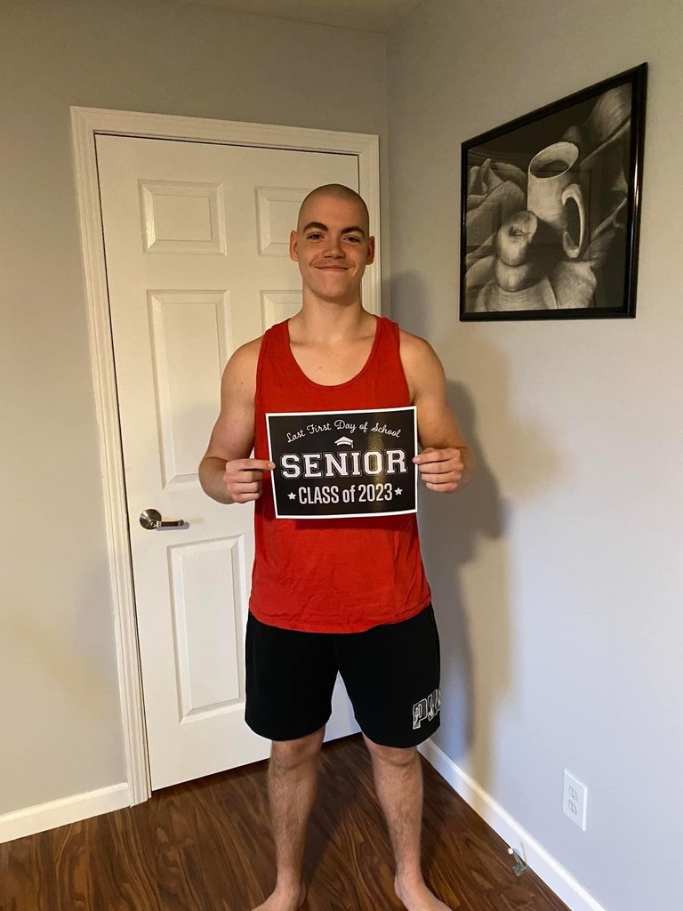A student in a red muscle shirt, and black jeans holds a black sign with white letters that reads "Last first day of school Seniors Class of 2023"