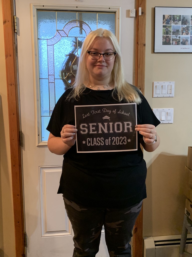 A student in a black shirt and black  pants holds a black sign with white letters that reads "Last first day of school Seniors Class of 2023"