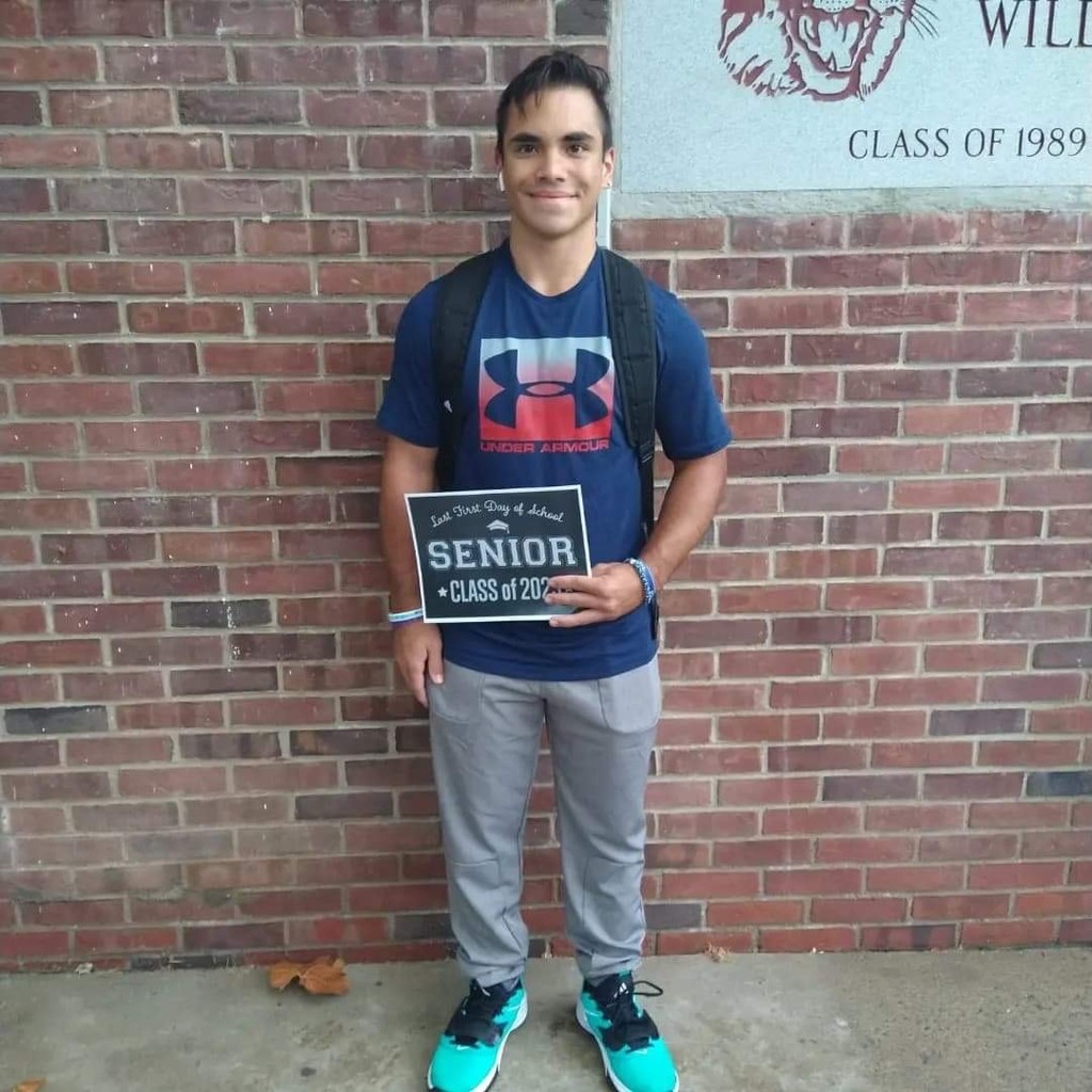A student in a dark blue shirt with a white and red design and gray pants holds a black sign with white letters that reads "Last first day of school Seniors Class of 2023"
