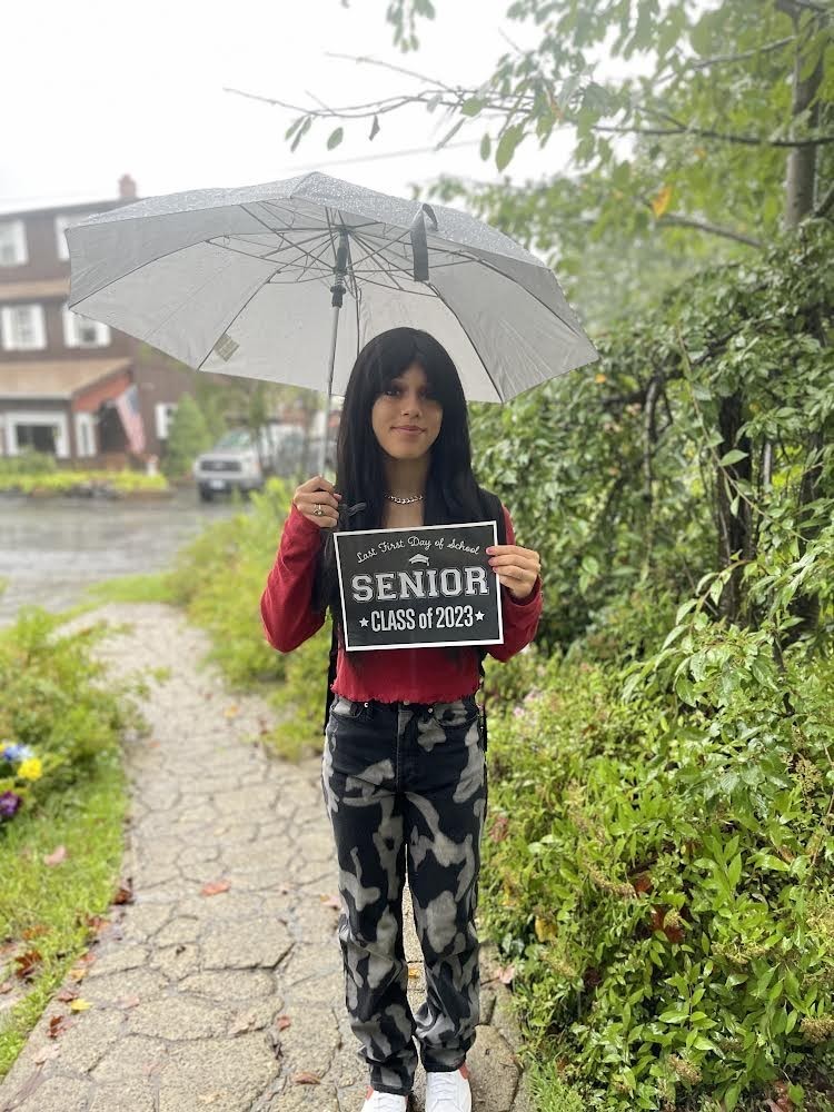 A student in a maroon  shirt and black, gray and white patterned pants,  holds an umbrella and a black sign with white letters that reads "Last first day of school Seniors Class of 2023"