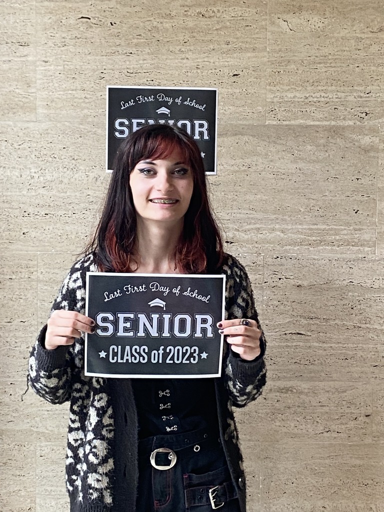 A student in a black shirt, black and white sweater and black pants holds a black sign with white letters that reads "Last first day of school Seniors Class of 2023"