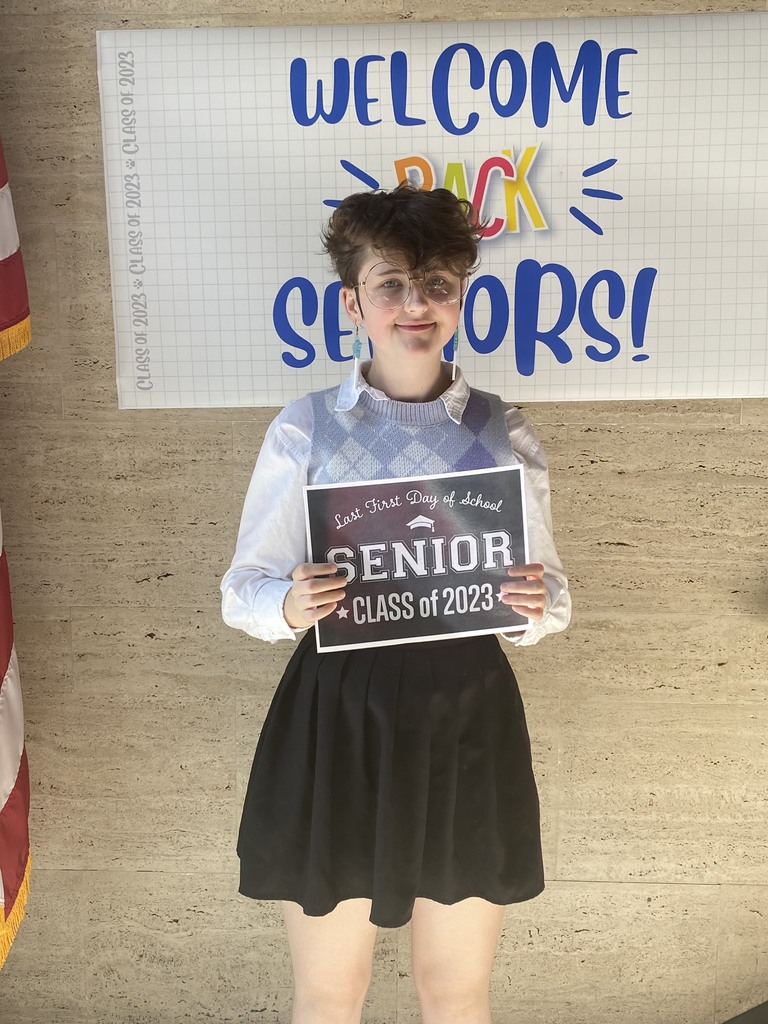 A student in white collared shirt and sweater vest and a black skirt holds a black sign with white letters that reads "Last first day of school Seniors Class of 2023"