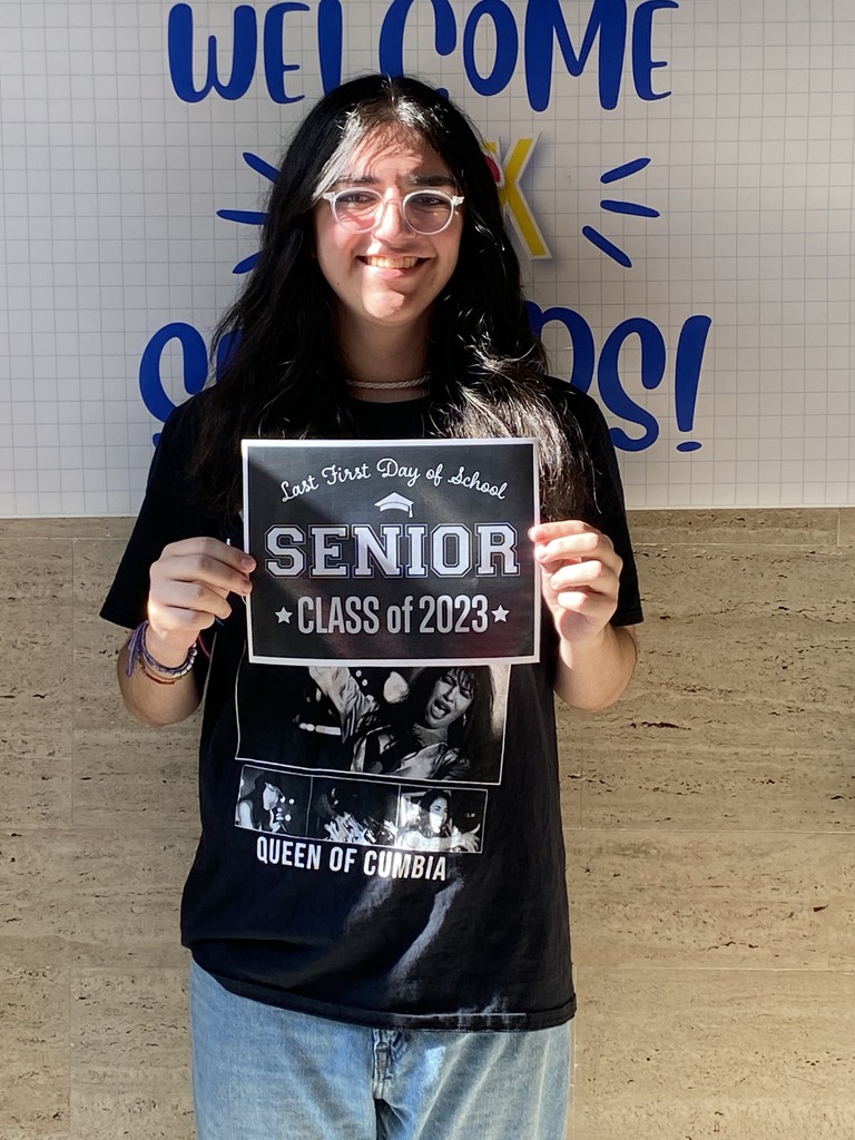A student in a black shirt and jeans holds a black sign with white letters that reads "Last first day of school Seniors Class of 2023"