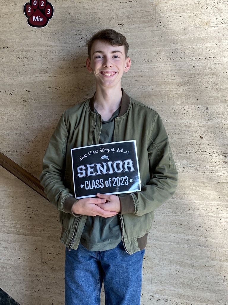 A student in a green longsleeved shirt and jeans holds a black sign with white letters that reads "Last first day of school Seniors Class of 2023"