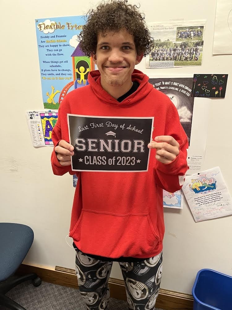 A student in a red hooded sweatshirt and black and white patterned pants holds a black sign with white letters that reads "Last first day of school Seniors Class of 2023"
