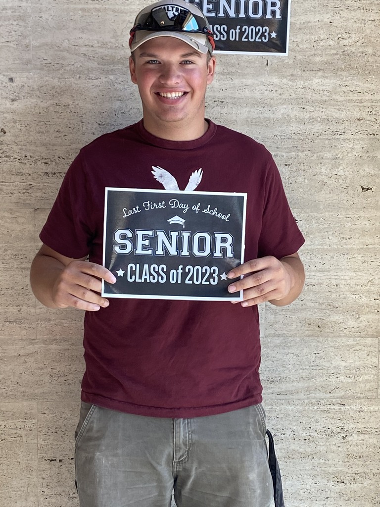A student in a maroon shirt, gray pants and a cap holds a black sign with white letters that reads "Last first day of school Seniors Class of 2023"