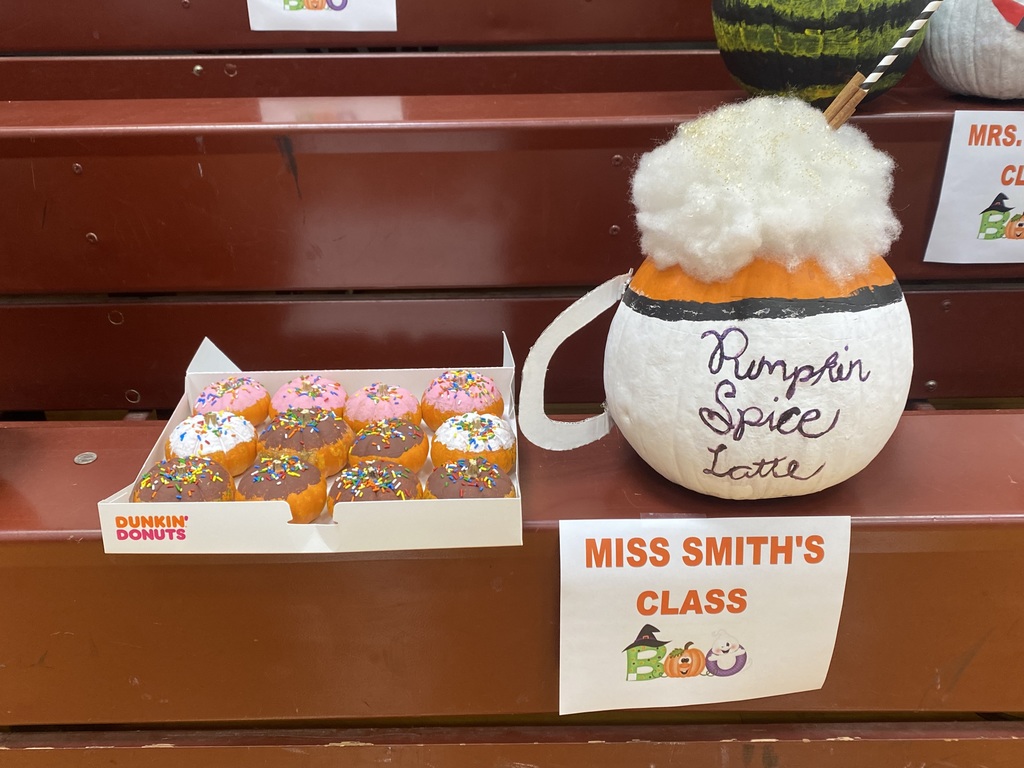 A pumpkin decorated as a cup of coffee sits next to a box of doughnuts with a sign below that reads Miss Smith's Class