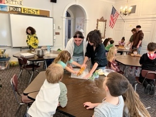 Two older students  help a younger student roll dough as other younger students watch
