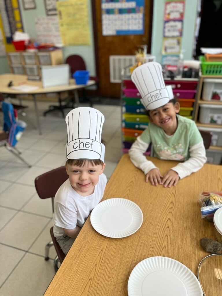 Two students sit at a table while wearing chef's hats