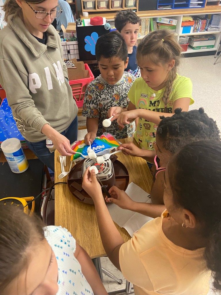 Students, with the help of an adult, roast marshmallows on forks over a s'mores maker.