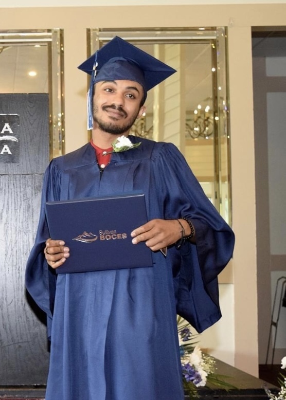 A student in a blue cap and gown holds a diploma