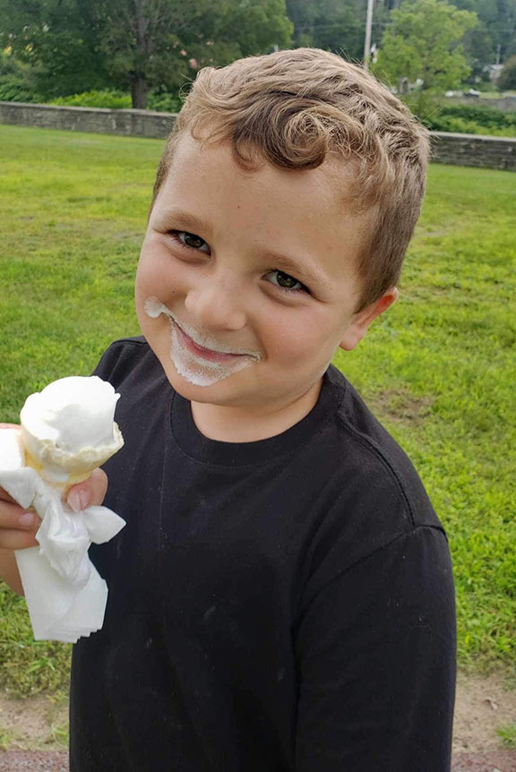 A student holds an ice cream cone and smiles for the camera with icecream on his face.