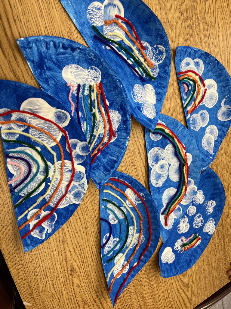 Half paper plates painted as blue skies with clouds and colored strings as rainbows are displayed on a table.