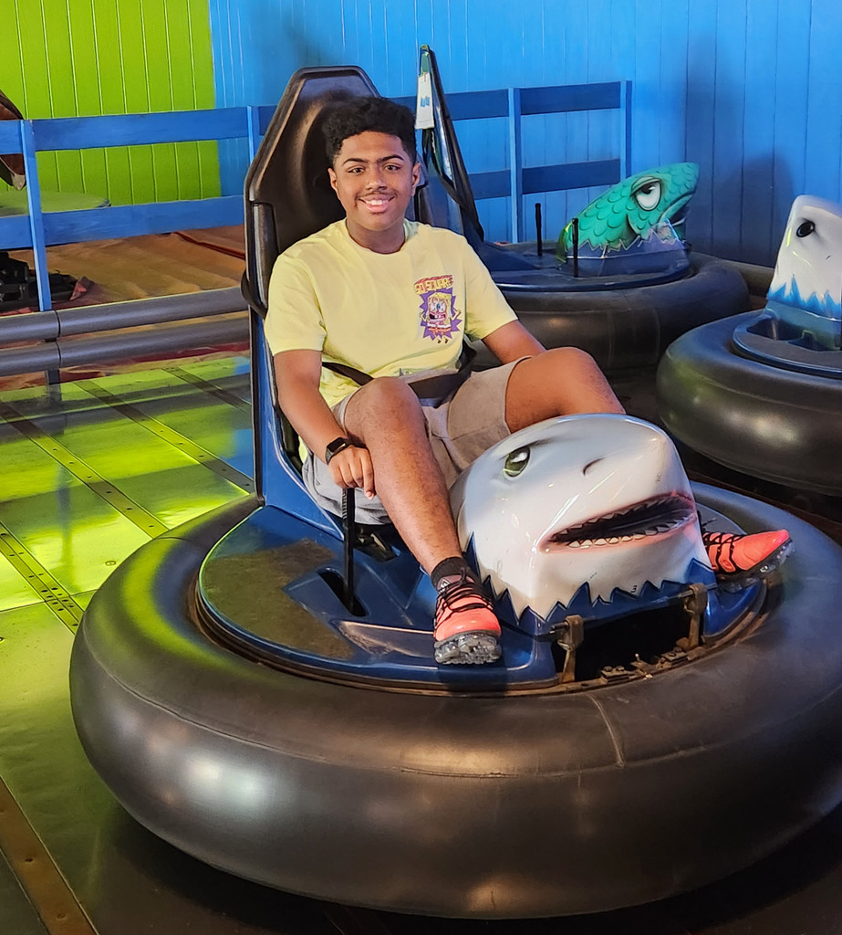 A student smiles while seated on a bumper car that has a shark head on it.