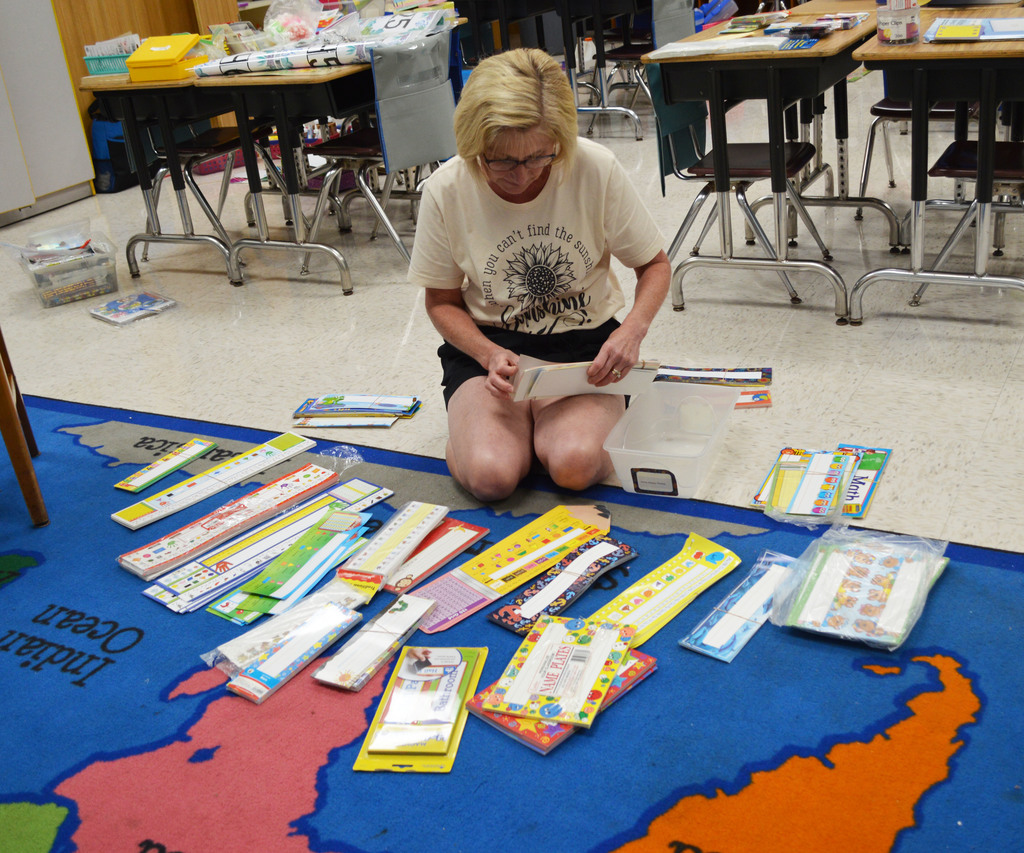 A teacher kneels next to a rug with a picture of a world map sorting paper items.