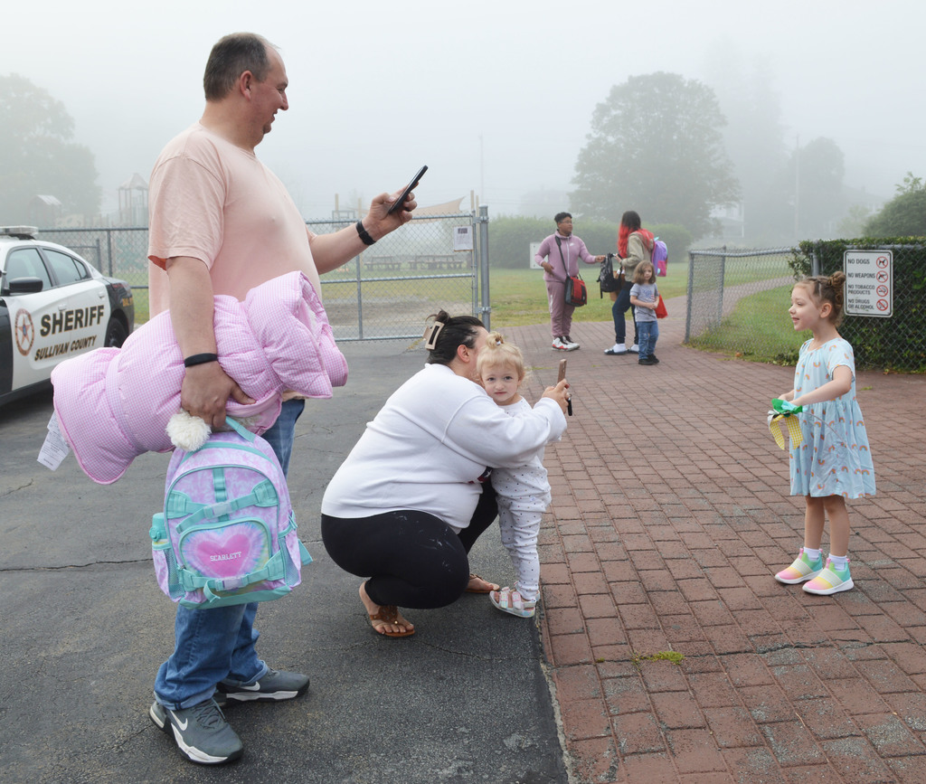 A young student poses for photos as two adults, one holding a blanket and backpack and the other squatting and embracing a toddler, take photos with their phones.
