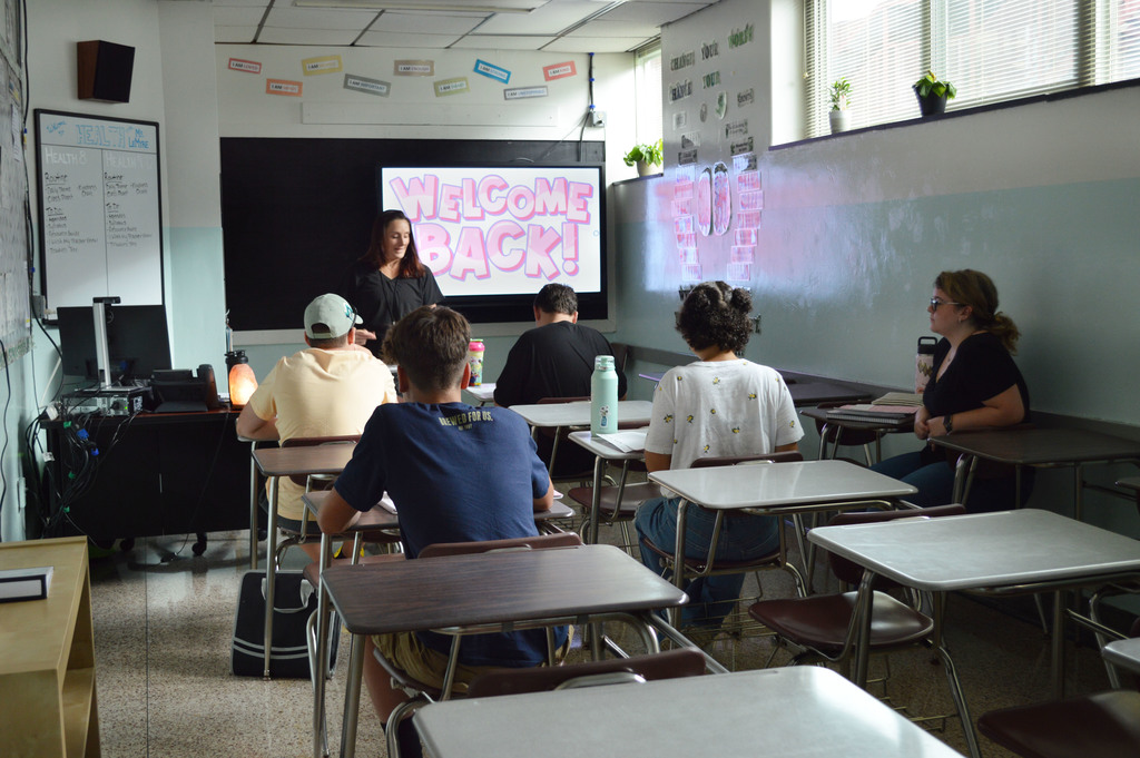 A teacher with Welcome Back on a screen behind her talks to a class of five students seated at desks