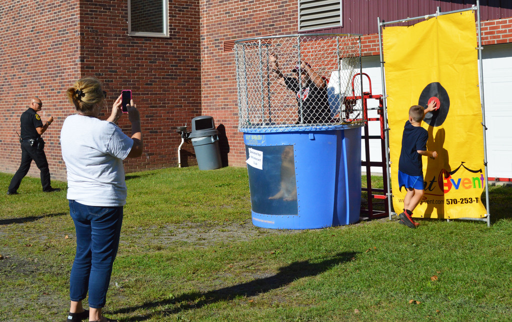 A student presses a button on a dunk tank as and adult falls in and another adult takes a photo with a phone.