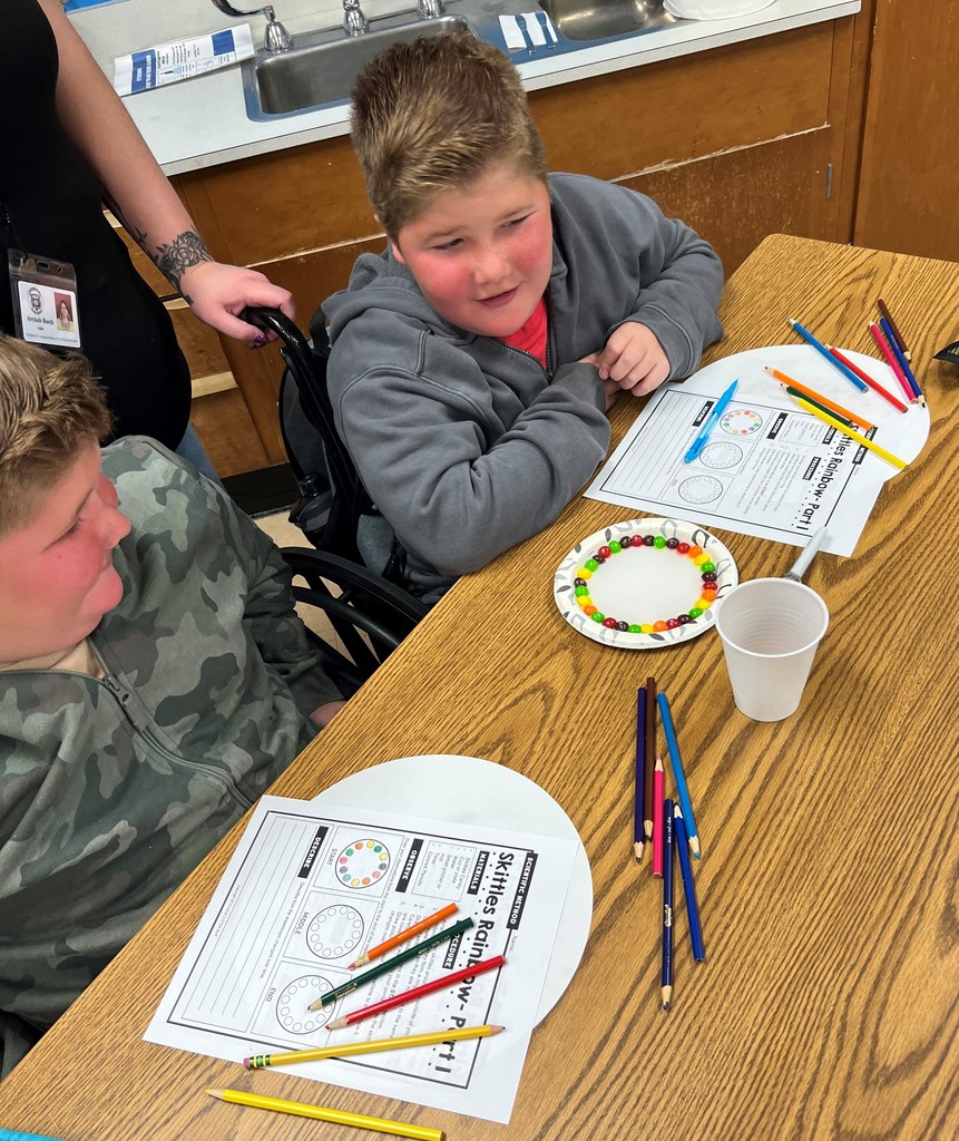 Two students sit at a desk with colored pencils, worksheets, a small plate with Skittles in a circle and a small cup as an adult stand behind them.