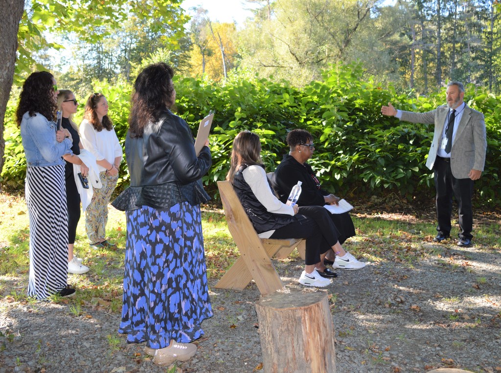 a man gestures as  two people sit on a bench and other stand in an outdoor classroom.