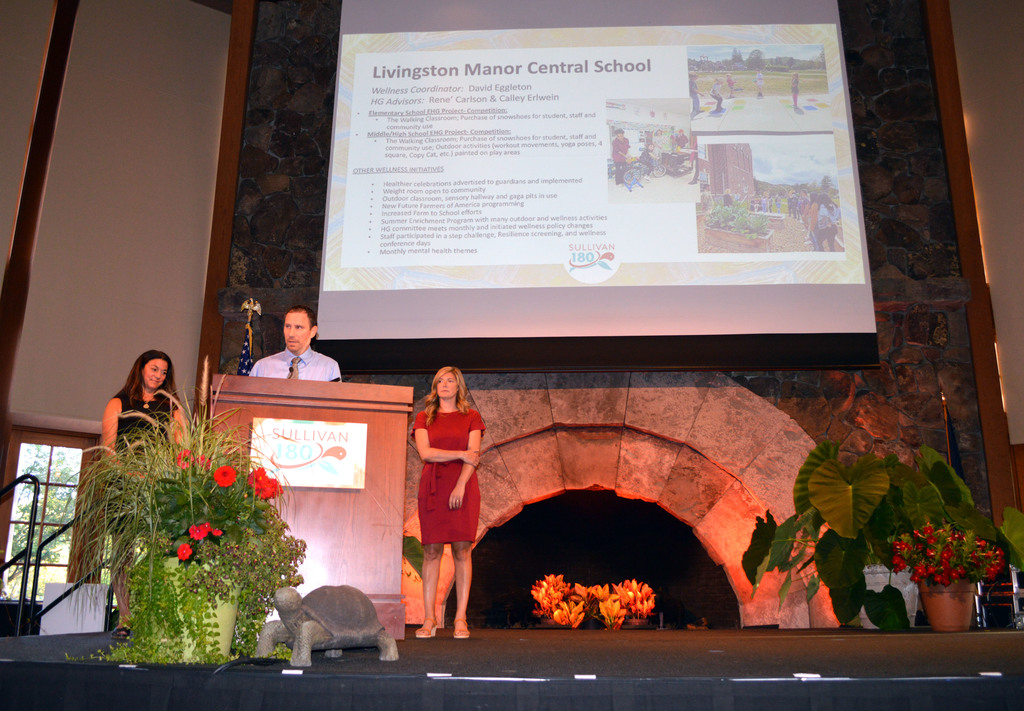 A person speaks at a podium flanked by two other people and a screen behind them displays a list of health and wellness projects done by LMCS