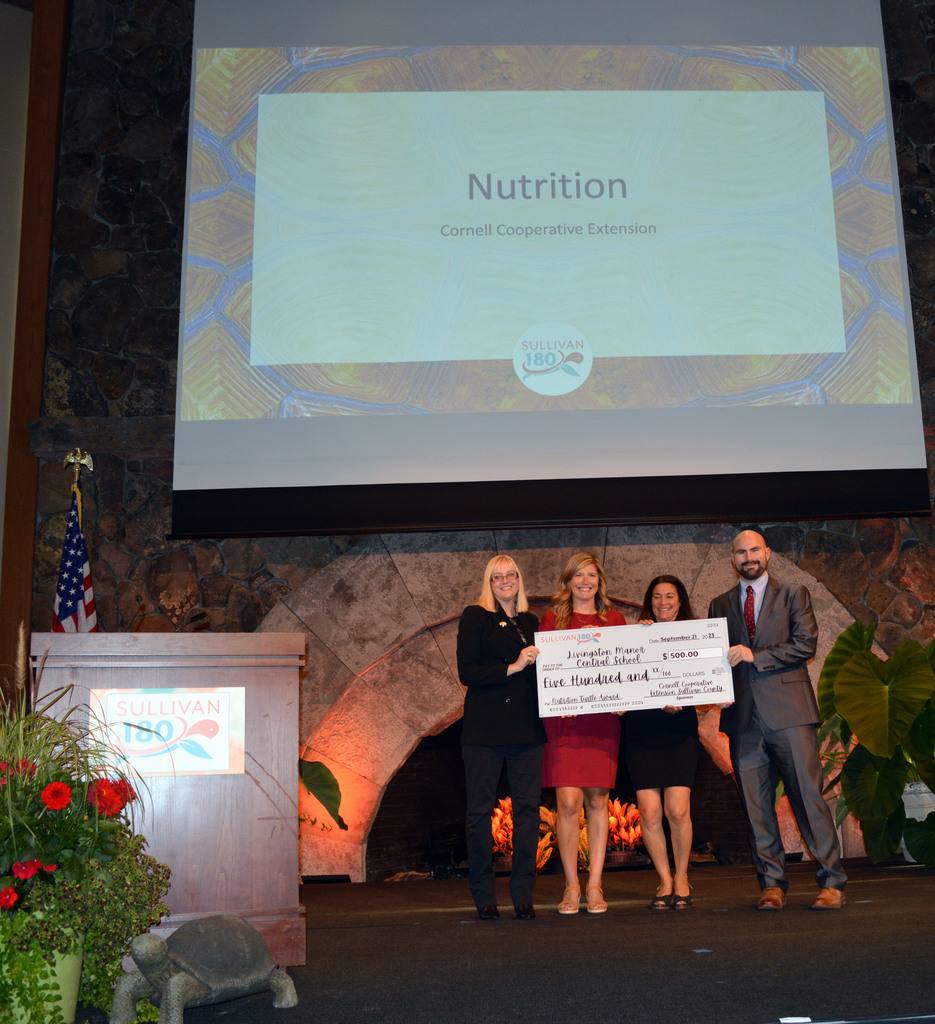Four people hold a big check on  a stage with a podium at left that has the Sullivan 180 logo on it and a screen behind them reads Nutrition Cornell Cooperative Extension