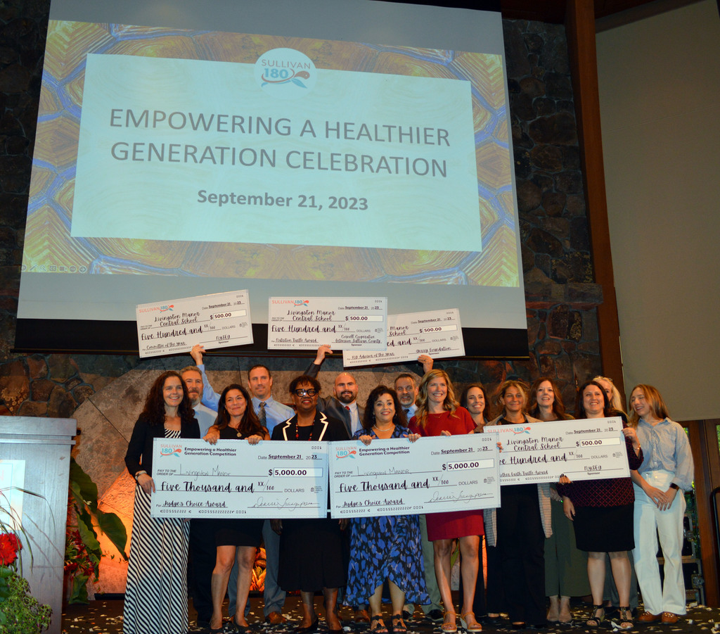 People stand on a stage hold 6 large checks with "Empowering a Healthier Generation Celebration, September 21, 2023 in the background