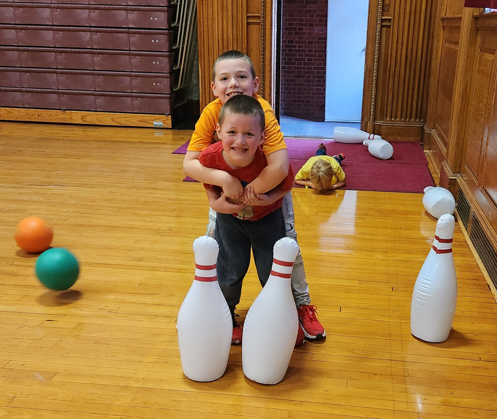 A student hugs another from behind  as bowling pins and balls and a student laying on the floor are around them