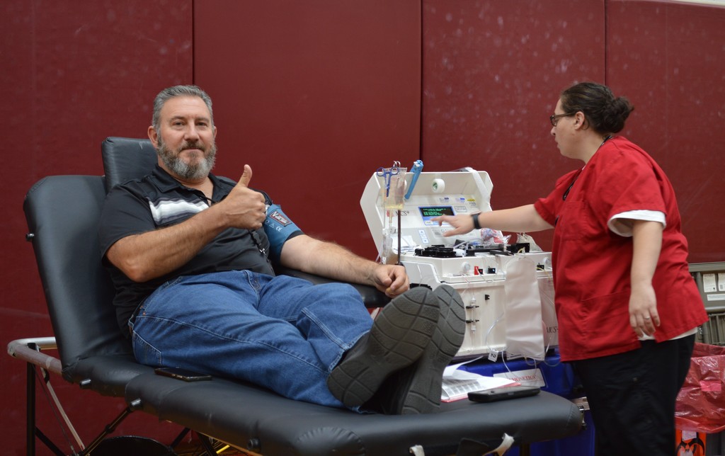 A man sitting on a cot gives a thumbs up as a person near him sets up a machine for blood donation